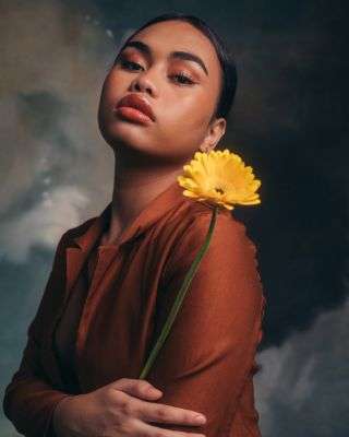 young person wearing their hair back, a rusty colored shirt, and holding a yellow daisy